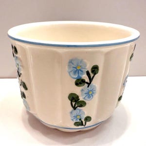 Peut inclure: Pot de fleurs en céramique crème avec un design cannelé. Il est orné de motifs floraux bleus et de feuilles vertes. Une fine ligne bleue souligne le bord. Un récipient décoratif pour plantes.