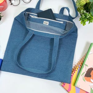 May include: A blue denim tote bag with a zippered pocket on the inside. The bag has two long straps and is lying flat on a white surface.