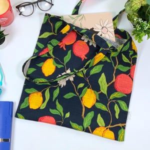 Puede incluir: Una bolsa de tela negra con estampado floral de limones, granadas y hojas verdes. La bolsa tiene dos asas y está hecha de un tejido resistente.