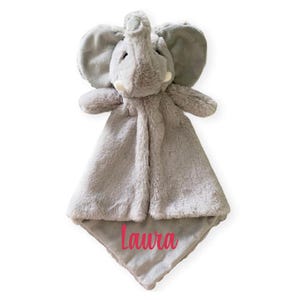 Puede incluir: Una manta de seguridad de peluche gris con forma de elefante, que presenta una cabeza de elefante con orejas grandes y colmillos peque&ntilde;os. El cuerpo de la manta es de tela suave y tiene el nombre Laura bordado en rosa.