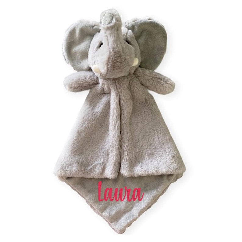 Puede incluir: Una manta de seguridad de peluche gris con forma de elefante, que presenta una cabeza de elefante con orejas grandes y colmillos peque&ntilde;os. El cuerpo de la manta es de tela suave y tiene el nombre Laura bordado en rosa.