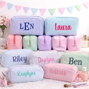 May include: A collection of personalized cosmetic bags in various pastel colors, including blue, pink, green, and lavender. Each bag features a name embroidered in a contrasting color. The bags are striped and have a zipper closure.