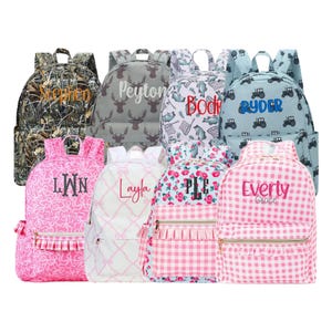 May include: A collection of personalized backpacks in various patterns and colors. Designs include camouflage, deer, fish, tractors, and floral prints. Each backpack features a name embroidered in a contrasting color.