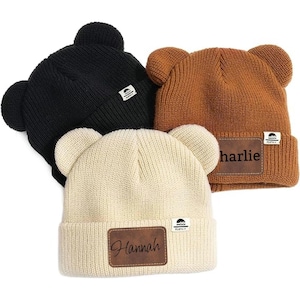 Peut inclure: Trois bonnets en tricot avec des oreilles d'ours. Les bonnets sont de couleur marron, noir et crème. Chaque bonnet a un patch en cuir avec un nom dessus. Les noms sont "Charlie", "Hannah" et "[nom illisible]".