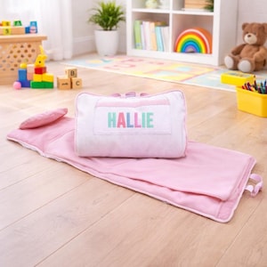 May include: A pink nap mat set with a pillow and attached blanket. The pillow is white with the name "HALLIE" in pastel colors. The mat has a rolled-up design with a handle for easy carrying. Perfect for toddlers and young children.