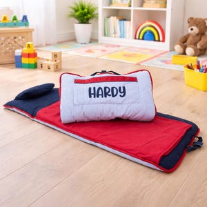 May include: A red and navy blue nap mat with a matching pillow. The pillow is white with blue stripes and the name "HARDY" in navy blue. The mat is designed for children and has a handle for easy carrying.