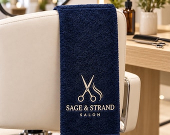 Puede incluir: Una toalla rectangular de color azul marino con un acabado de tela de rizo texturizada. La toalla presenta un logotipo bordado en dorado de unas tijeras y un mechón de pelo estilizado sobre el texto SAGE & STRAND SALON.