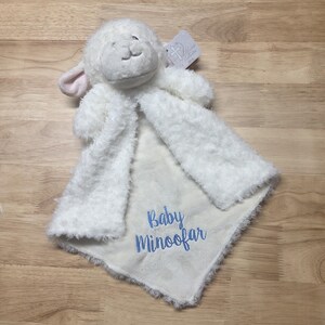 Puede incluir: Una manta de seguridad de peluche blanco con forma de cordero y textura suave y esponjosa. El centro presenta el texto bordado en azul Baby Minoofar. La cabeza del cordero tiene orejas forradas en rosa y una peque&ntilde;a etiqueta.