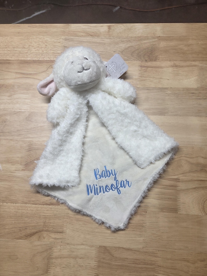 Puede incluir: Una manta de seguridad de peluche blanco con forma de cordero y textura suave y esponjosa. El centro presenta el texto bordado en azul Baby Minoofar. La cabeza del cordero tiene orejas forradas en rosa y una peque&ntilde;a etiqueta.
