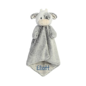 Puede incluir: Una manta de seguridad de peluche con forma de vaca y textura de piel sint&eacute;tica gris y blanca. La cabeza de la vaca tiene rasgos faciales bordados y una mancha sobre un ojo. El nombre Elliott est&aacute; bordado en hilo azul en la esquina.