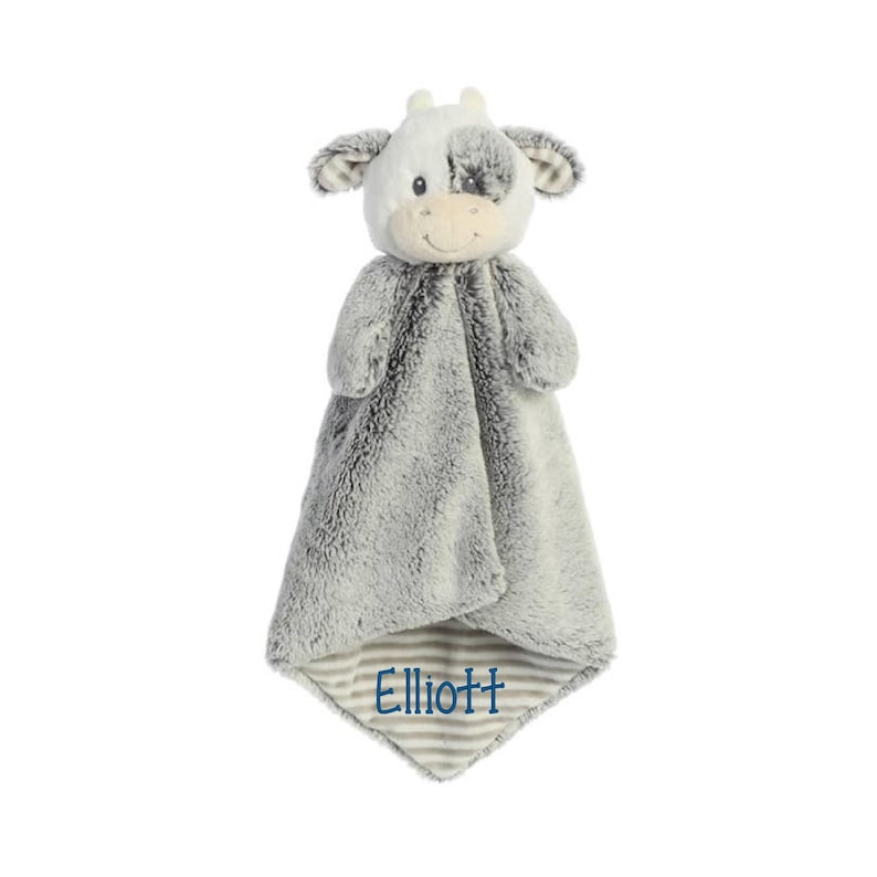 Puede incluir: Una manta de seguridad de peluche con forma de vaca y textura de piel sint&eacute;tica gris y blanca. La cabeza de la vaca tiene rasgos faciales bordados y una mancha sobre un ojo. El nombre Elliott est&aacute; bordado en hilo azul en la esquina.