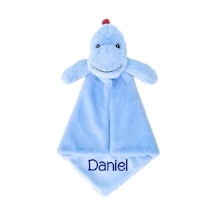 Puede incluir: Una manta de seguridad de felpa azul claro con una cabeza de dinosaurio de peluche y una peque&ntilde;a cresta roja en la parte superior. El cuerpo de tela suave y difusa tiene forma de diamante y lleva el nombre Daniel bordado en hilo azul oscuro.