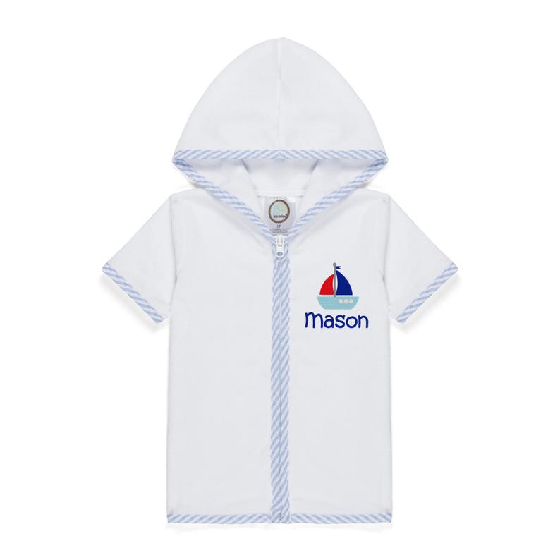 Peut inclure: Peignoir blanc &agrave; capuche et manches courtes, bord&eacute; de rayures bleues et blanches. Motif de bateau &agrave; voile rouge, bleu et blanc, avec le nom "Mason" brod&eacute; en bleu marine. Fermeture &eacute;clair int&eacute;grale.