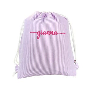 May include: A drawstring bag with a vertical striped pattern in lavender and white. The bag features the name "gianna" in a cursive, magenta font across the front. White drawstrings are visible at the top.