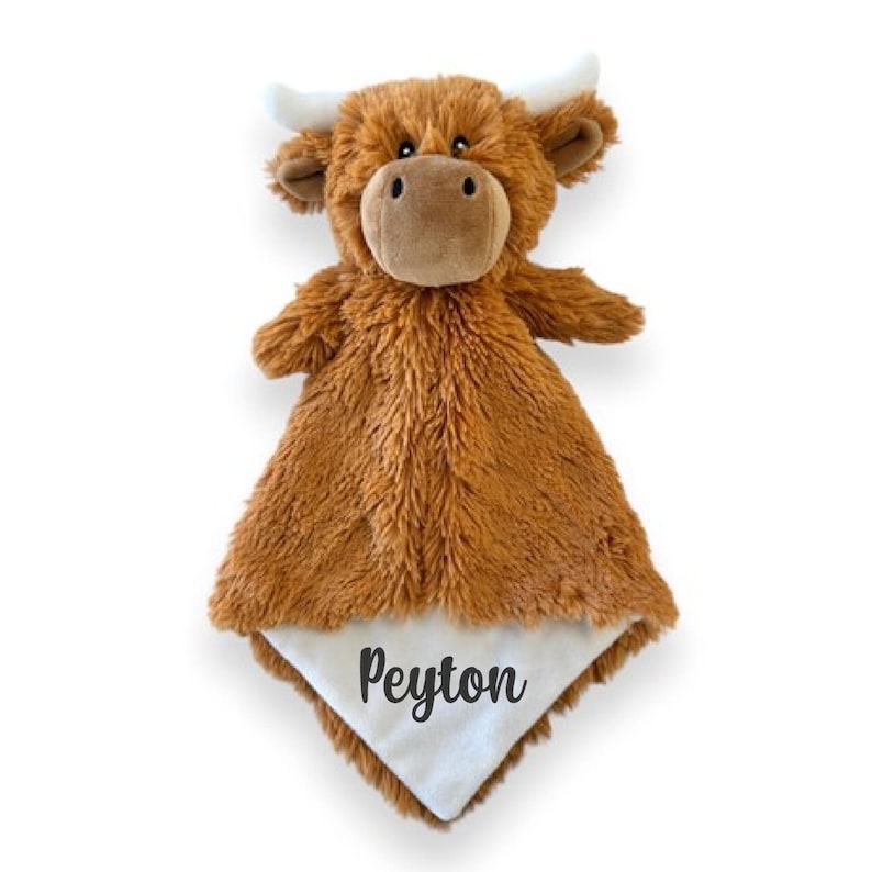 Puede incluir: Una manta de seguridad de peluche con forma de vaca Highland, con textura marr&oacute;n peluda, cuernos blancos y un parche triangular blanco liso en la esquina bordado con el nombre Peyton en una fuente cursiva gris oscuro.