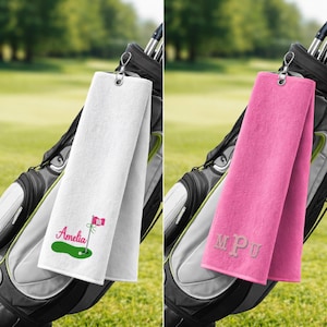 May include: Two golf towels hanging from a golf bag. One is white with a green golf course design and the name "Amelia" in pink. The other is pink with the initials "MPU" embroidered in a lighter pink.