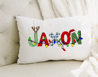 Puede incluir: Almohada blanca con el nombre Jaxon escrito en letras coloridas con un tema navideño. Las letras están decoradas con luces, cuadros y un gorro de Papá Noel.