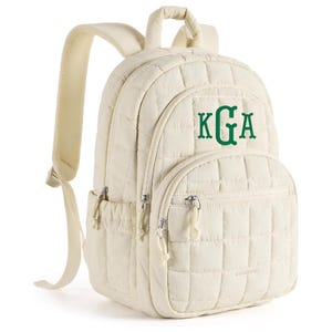 May include: A cream-colored quilted backpack featuring a grid pattern and a top handle. The front displays the embroidered green monogram letters KGA. It includes adjustable shoulder straps, side pockets, and silver-toned metal zipper pulls.