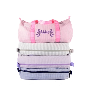 May include: A stack of four seersucker duffel bags. The top bag is pink and white with bright pink handles and the name Addie embroidered in purple. The other three bags are grey and white, lavender and white, and blue and white, each with a zipper.