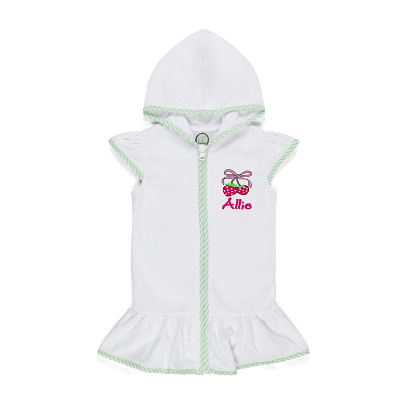 Peut inclure: Robe de b&eacute;b&eacute; blanche &agrave; capuche avec fermeture &eacute;clair et ourlet &agrave; volants. La robe pr&eacute;sente une bordure verte et un motif de cerises avec le nom "Allie" brod&eacute; en rose.