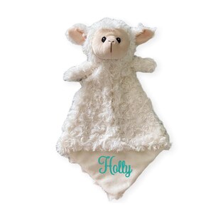 Puede incluir: Una manta de seguridad de peluche con forma de cordero blanco, con un cuerpo de textura rizada suave y una cara lisa de color claro. En la esquina inferior de la manta aparece el nombre Holly bordado en letra cursiva turquesa.