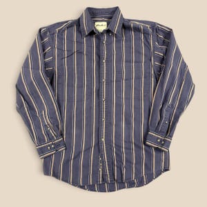 May include: A long-sleeve button-down shirt in a muted blue with vertical stripes in tan and dark blue. The shirt features a button-down collar, two chest pockets, and a front button placket. The interior neck label reads Eddie Bauer.