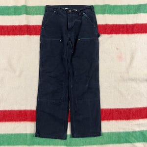 May include: Dark navy blue work pants featuring a straight-leg cut, a button-fly closure, and reinforced double-layer fabric panels on the front of the thighs. These pants include metal rivets at the pocket corners and a standard waistband.