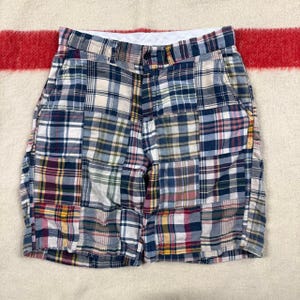 May include: A pair of shorts featuring a patchwork design composed of various plaid fabric squares in shades of blue, white, red, yellow, and green. The shorts have a button closure at the waist, a zipper fly, and two side pockets.