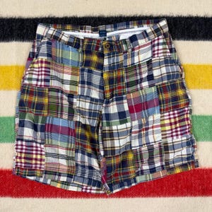 May include: A pair of shorts featuring a patchwork design composed of various plaid fabric squares in shades of blue, red, yellow, green, and white. The shorts include a waistband with a button closure, a front zipper fly, and side pockets.