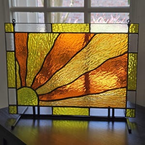 May include: Stained glass panel featuring a sunburst design in shades of yellow and orange. The glass is textured, with black lines separating the colors. The panel is rectangular and mounted on a black metal stand.