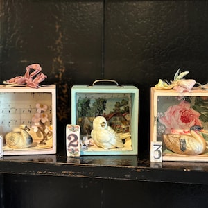 May include: Three square wooden shadow boxes featuring bird figurines. Box one has a swan and sheet music. Box two is light blue with a white bird and landscape print. Box three is light pink with a duck and floral wallpaper. Each has a numbered tag.
