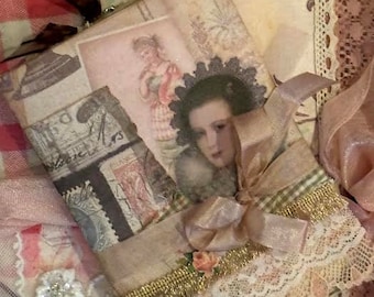 Puede incluir: Un diario de estilo vintage con una cinta rosa y dorada atada alrededor. El diario está decorado con encaje, flores de papel y sellos vintage. El diario es una excelente manera de registrar tus pensamientos y recuerdos.