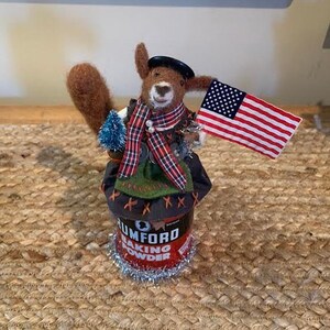 May include: A brown and white furry animal wearing a red and black plaid scarf and a black hat, holding a small American flag. The animal is standing on a red and white can that says "Rumford Baking Powder".
