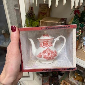 May include: A white porcelain teapot with a red and white floral design sits in a small wooden box with a red trim. The teapot has a spout and a handle. The box is open and the teapot is visible inside.