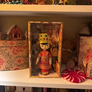 May include: A vintage China doll in a decorative box. The doll wears a red and gold outfit with a jeweled headdress. The box has floral and butterfly imagery. The doll is displayed on a white shelf with other vintage items.