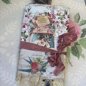 May include: A vintage-style journal with a floral pattern and a ribbon tie. The journal features images of vintage perfume labels and a bird with flowers. A lace trim and a decorative pin with beads add detail.