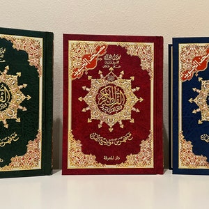 May include: Three hardcover books with ornate gold and red accents. The books are in green, red, and blue, each featuring Arabic script and a central starburst design. The text includes "Dar Al-Ma'arifa" and "Quran".