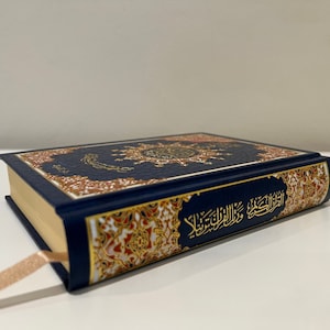 May include: A closed, navy blue book with gold gilded edges and a decorative cover featuring ornate gold and red patterns. The book has a light brown ribbon bookmark. The cover has Arabic script.