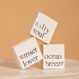 May include: Three white wooden blocks with black text. The top block reads "salty soul", the middle block reads "sunset lover", and the bottom block reads "ocean breeze".