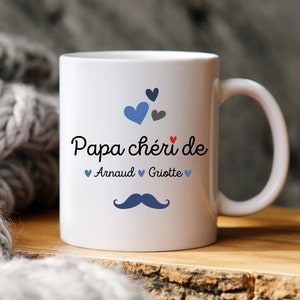 Peut inclure: Mug en céramique blanche avec un motif de cœurs bleus et rouges. Le texte "Papa chéri de Arnaud Griotte" est imprimé sur le mug.