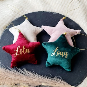 May include: Four plush star ornaments in white, pink, burgundy, and emerald green. Each star has a gold string for hanging and a name in gold script. The burgundy star says "Lola" and the emerald green star says "Louis".