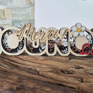 Peut inclure: Panneau décoratif en bois avec les mots "Nounou Merci Louis" dans un design découpé au laser en couches. Le panneau présente une marguerite et des motifs floraux. Le nom "Louis" est en rouge. Le panneau est sur une surface en bois.