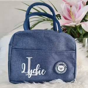 以下が含まれることがあります： 白いクマのロゴと「Lunch Bag Design by Livework」の文字が入った青いデニムのランチバッグ。バッグには「Lydie」という名前が銀色のグリッターでパーソナライズされています。