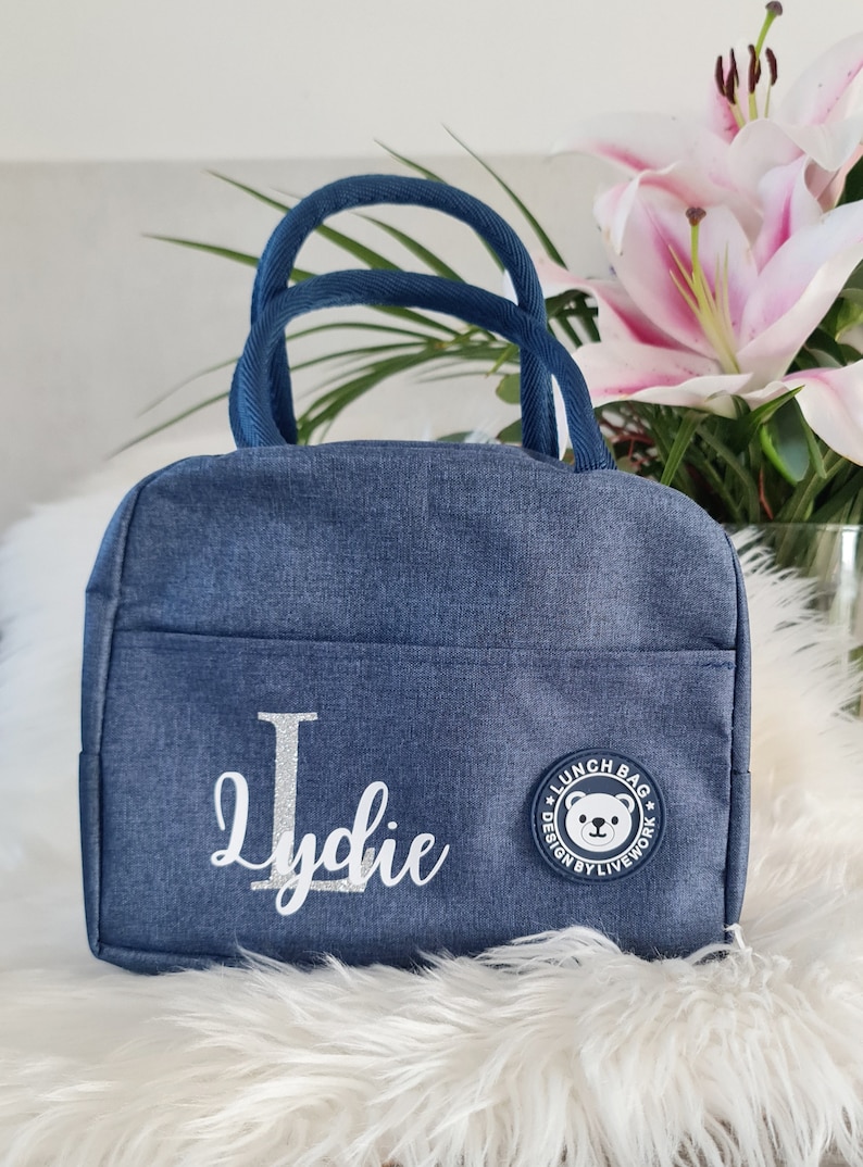 以下が含まれることがあります： 白いクマのロゴと「Lunch Bag Design by Livework」の文字が入った青いデニムのランチバッグ。バッグには「Lydie」という名前が銀色のグリッターでパーソナライズされています。