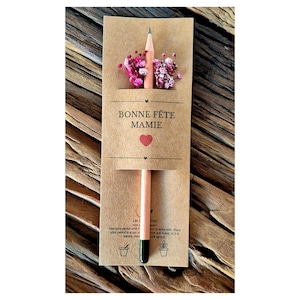 Peut inclure: Un crayon rose clair avec une pointe noire et un petit bouquet de fleurs roses, présenté sur une carte marron. La carte porte l'inscription "BONNE FÊTE MAMIE" avec un cœur rouge et de petites icônes.