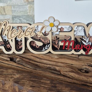 May include: Wooden decorative sign with the word "Merci!" in cursive, followed by "Margot" in red, layered with floral and butterfly cutouts. A white daisy with a yellow center is placed above the "S" in "Merci!"