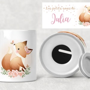 May include: White ceramic piggy bank featuring a watercolor illustration of a fox with floral accents and a butterfly. The fox is brown and white with pink flowers. The bank has a coin slot and a removable bottom. Text on the top reads "Les petits sous de Julia".