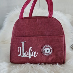 以下が含まれることがあります： 白熊のロゴと「Lunch Bag Design by Livework」の文字が入った、ワインレッドのランチバッグ。バッグには「Lola」という名前がシルバーのグリッターでパーソナライズされています。
