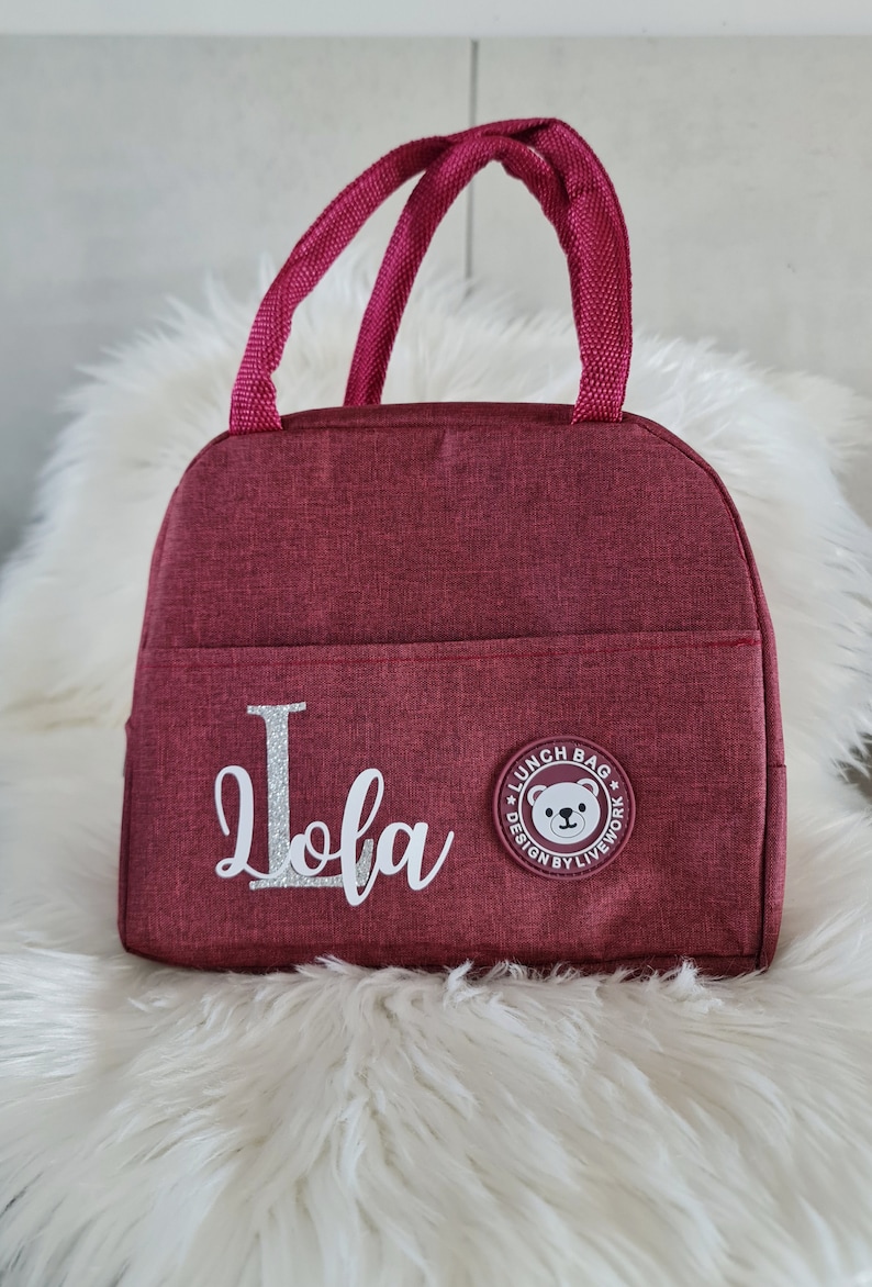 以下が含まれることがあります： 白熊のロゴと「Lunch Bag Design by Livework」の文字が入った、ワインレッドのランチバッグ。バッグには「Lola」という名前がシルバーのグリッターでパーソナライズされています。