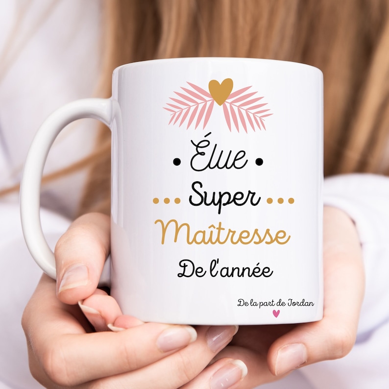 May include: White ceramic mug with a pink and gold design. The text on the mug reads "&Eacute;lue Super Maitresse De l'ann&eacute;e De la part de Jordan".